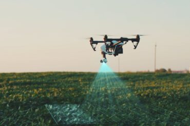 A white agricultural drone flies over the farm fields, scanning and analyzing crops, under a clear sky.
