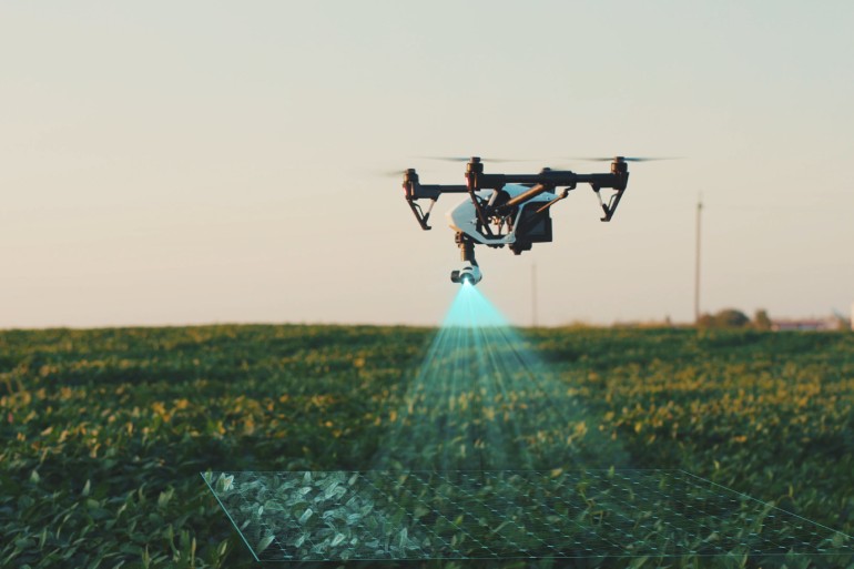 A white agricultural drone flies over the farm fields, scanning and analyzing crops, under a clear sky.