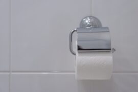 A close-up view shows a silver wall-mounted toilet paper holder on a white-tiled wall. The holder contains toilet paper.