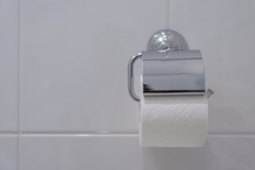 A close-up view shows a silver wall-mounted toilet paper holder on a white-tiled wall. The holder contains toilet paper.