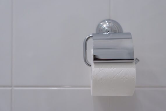 A close-up view shows a silver wall-mounted toilet paper holder on a white-tiled wall. The holder contains toilet paper.