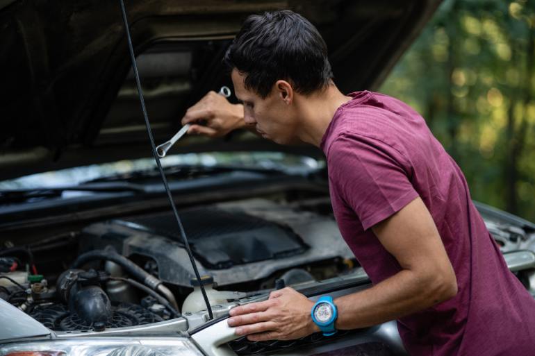 A brown-haired man in a maroon T-shirt and blue watch stands in front of a car engine with a wrench in his hand.