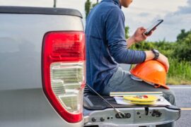 A man is sitting on the back of a pickup truck. He’s holding an orange hard hat in one hand and a phone in the other.