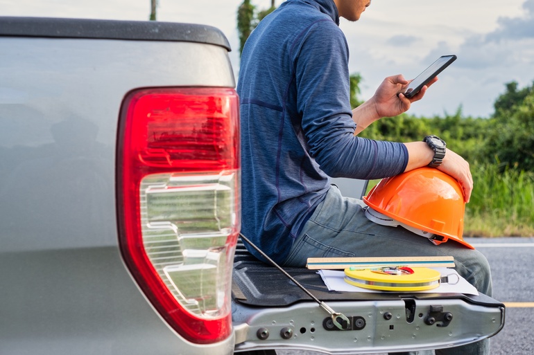 A man is sitting on the back of a pickup truck. He’s holding an orange hard hat in one hand and a phone in the other.