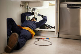 A man in blue coveralls is laying down under a sink. He’s using a tool on the plumbing underneath the sink.
