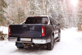 A large blue truck is driving through a small road in a forest that is covered with snow. The truck also has snow on it.