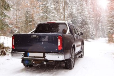 A large blue truck is driving through a small road in a forest that is covered with snow. The truck also has snow on it.