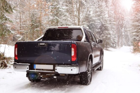 A large blue truck is driving through a small road in a forest that is covered with snow. The truck also has snow on it.