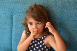 A young girl with brown hair sucks her thumb and plays with her hair. She is wearing a blue polka-dot top.