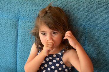 A young girl with brown hair sucks her thumb and plays with her hair. She is wearing a blue polka-dot top.