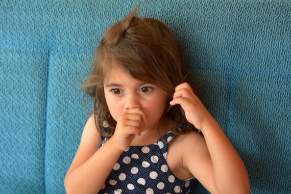 A young girl with brown hair sucks her thumb and plays with her hair. She is wearing a blue polka-dot top.