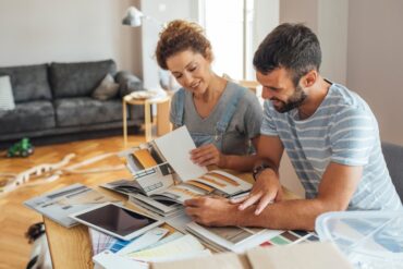 A man and a woman are sitting next to each other as they both look through a magazine that has pictures in it.