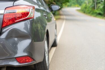 The rear of a gray car parked on the side of a clear road. The vehicle's back bumper has a large dent.