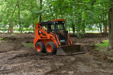 A skid steer sits in the middle of a clearing. There are trees in the background as well as a fence.