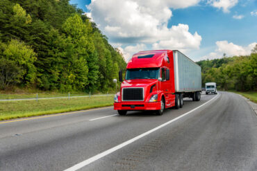 A semitruck drives along a two-lane street lined with dense foliage. The vehicle has a red tractor unit.