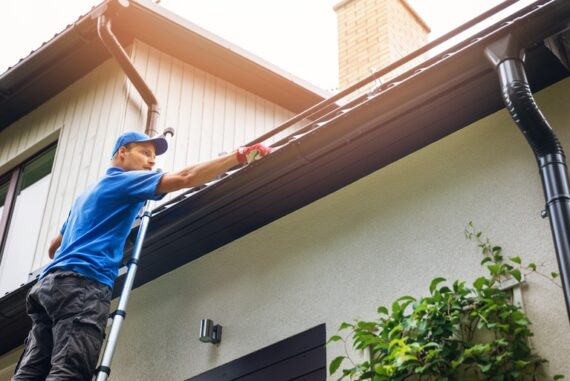A man in a blue uniform shirt and hat wears thick gloves while cleaning the gutters of a luxury home.