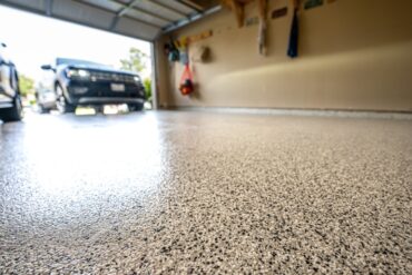 A garage floor with a fresh coating of epoxy and flakes. There are cars parked in the driveway behind the garage door.