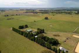 An aerial view of a large plot of rural land. Open fields of grass connect to a homestead with several buildings and trees.
