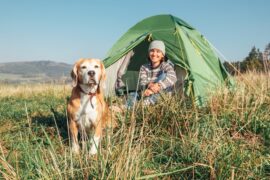 A woman in a sweater and a beanie smiles at her dog while sitting in front of a green tent outdoors during daytime.