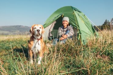 A woman in a sweater and a beanie smiles at her dog while sitting in front of a green tent outdoors during daytime.