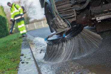 In the foreground is a street sweeping vehicle with a large brush cleaning the road, while in the background is a man standing in place.