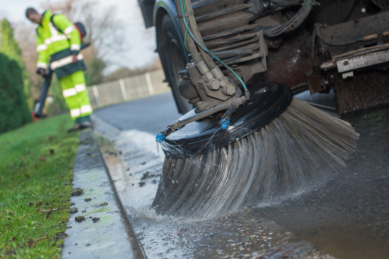 In the foreground is a street sweeping vehicle with a large brush cleaning the road, while in the background is a man standing in place.