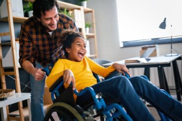 A daughter smiles and laughs as her dad tilts her wheelchair backward while they play. They are in a home office.