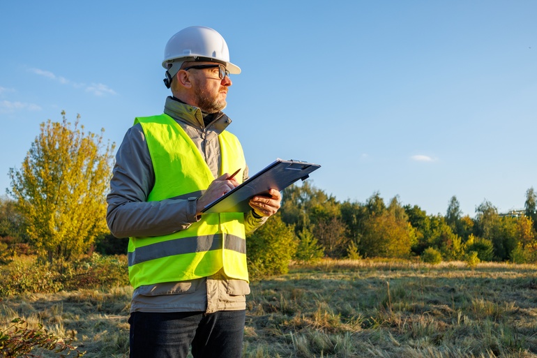 A man in a fluorescent green vest and protective glasses is standing in a field while he is holding a clipboard.