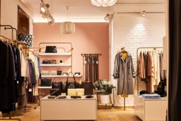 A boutique with one pink wall and one white brick wall. Racks of clothes and displays with wallets and purses fill the shop.