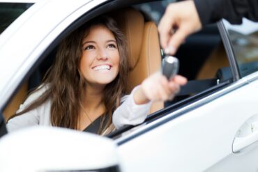 A woman with long brown hair is sitting in the driver’s seat of a car while someone hands her keys.