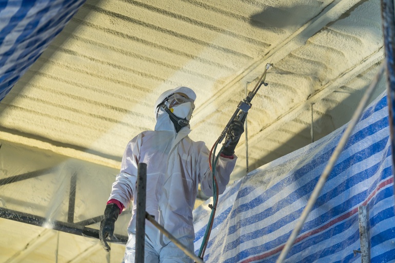 A technician wearing a protective suit and mask applies spray foam insulation on the ceiling of a warehouse.