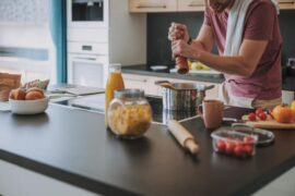 A man grinds pepper over a pot on a modern stovetop, with a pasta jar, tomatoes, and bread on the counter.