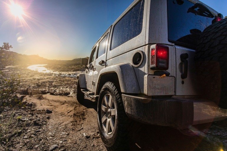 A white Jeep Wrangler drives through the mud on a rocky riverbank at sunset, with a spare tire carrier on its back end.