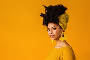 A woman posing in a yellow shirt, large yellow tassel earrings, and her unstyled afro held up with a yellow wrap.