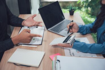 The hands of two businesspeople over a desk. There is a laptop and multiple papers on the desk below them.