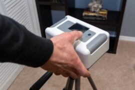 A close-up view shows a person pressing a gray button on a white, square device that sits on a black tripod.