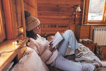 A woman in a knit hat reads a book on a bed inside a cozy wooden cabin, with string lights and a sunlit window nearby.