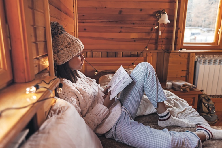 A woman in a knit hat reads a book on a bed inside a cozy wooden cabin, with string lights and a sunlit window nearby.