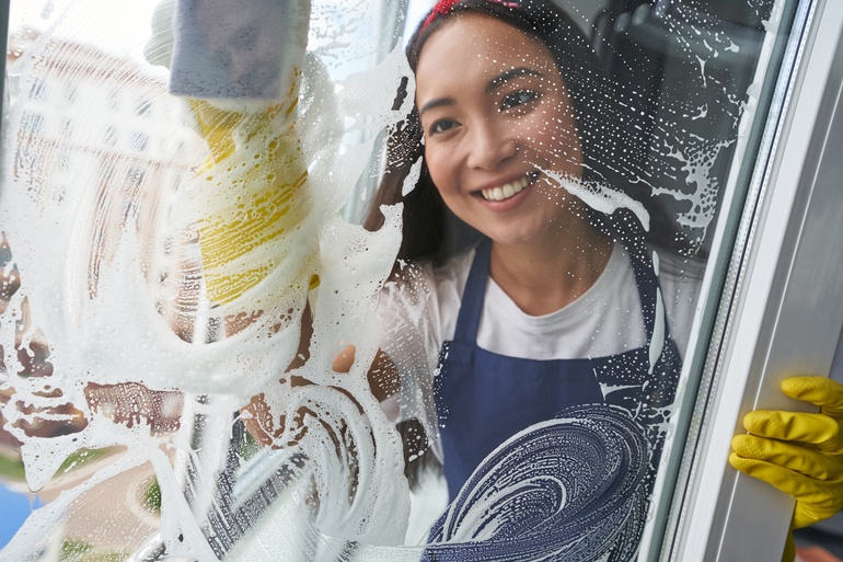 A woman smiles as she cleans off a window in her home. She wears yellow gloves and a blue apron as she cleans.