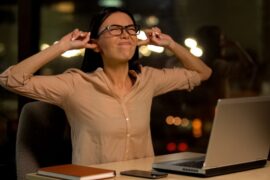 A woman at a desk covering her ears in frustration while working on a laptop at night in a softly lit office.