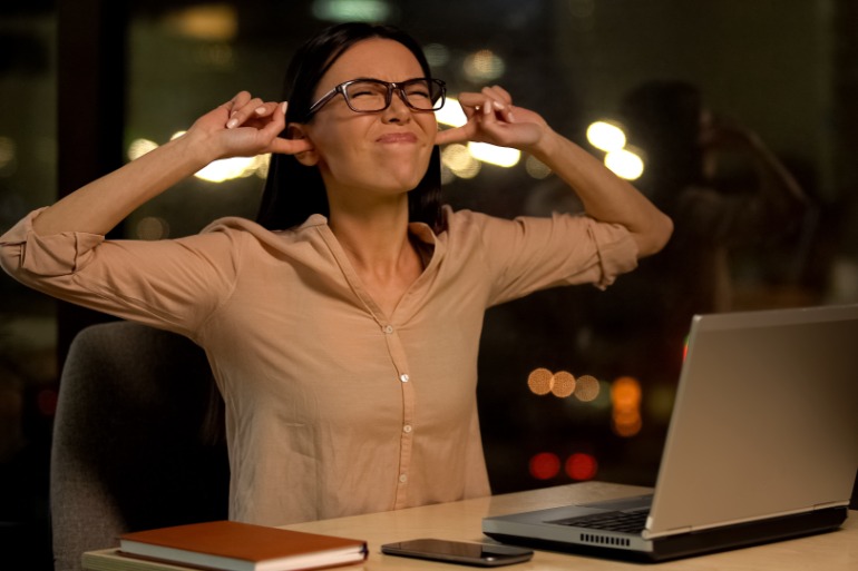 A woman at a desk covering her ears in frustration while working on a laptop at night in a softly lit office.
