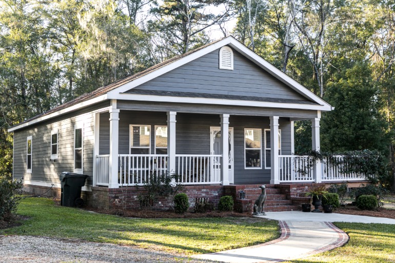 A gray single-story cottage with white trim and a covered front porch, brick steps, a wooded backyard, and a green lawn.