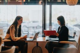 Two women facing each other across a small table. One has a laptop in front of her, the other holds a paper folder.