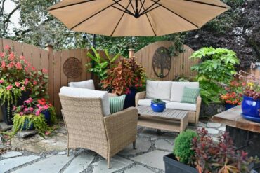 A cozy outdoor patio with wicker seating under an umbrella, surrounded by lush potted plants and wooden fence décor.