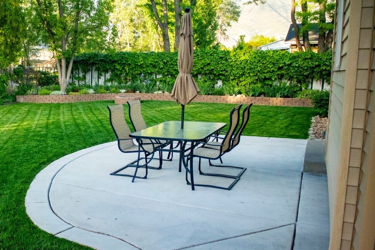 The backyard of a home with freshly cut grass, a concrete patio, and a glass table with four chairs and an umbrella.
