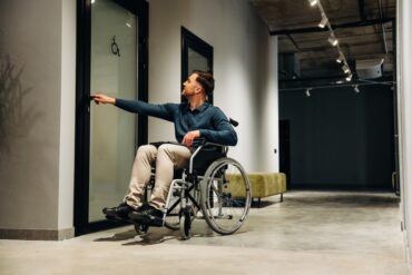 A young man in a wheelchair reaching for the door to another room. The door has a handicap symbol on it.