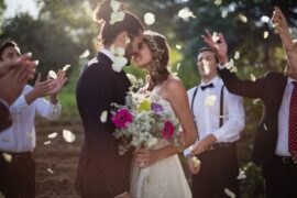 A bride and groom kiss while guests stand nearby and toss flower petals as the couple holds a bouquet outdoors.