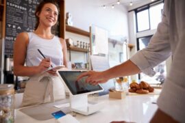 A customer uses their finger to touch the screen of an iPad kiosk while a smiling person stands behind the counter writing.