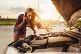 A woman wearing a flannel and shorts holds a phone up to her ear as she stands in front of her car with the hood open.