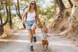 A woman and a young child walk hand in hand along a forest path, smiling during a sunny outdoor outing.